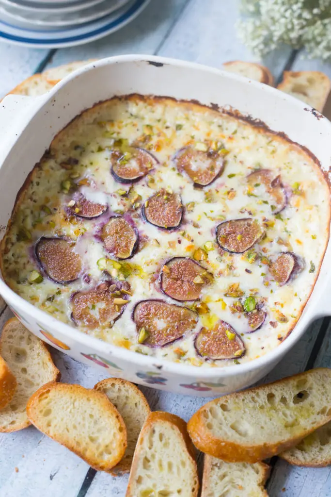 White dish with baked ricotta dip topped with figs and surrounded by sliced baguette
