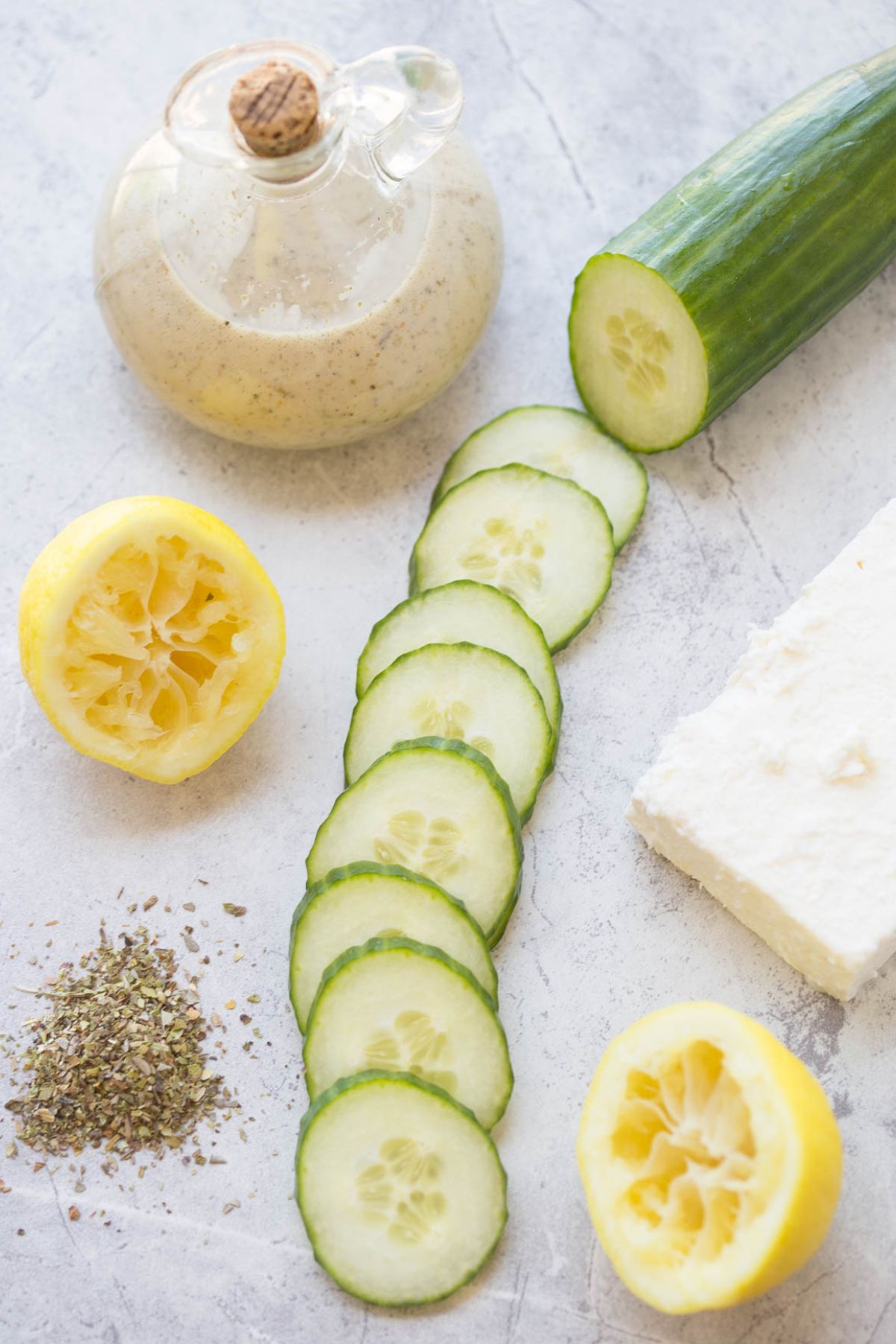 Ingredients for cucumber salad: sliced cucumbers in a line with halved lemons, a fresh garlic bulb, and a block of feta cheese, and a dressing jar.