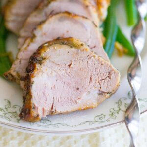 Garlic roasted pork tenderloin cut into rounds displayed on a white plate with green beans beside it.