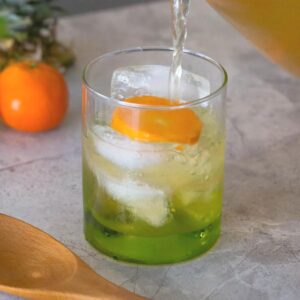 Pineapple orange green tea being poured into a green glass and garnished with a slice of orange