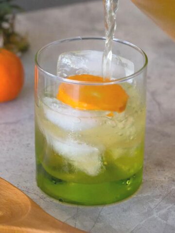 Pineapple orange green tea being poured into a green glass and garnished with a slice of orange