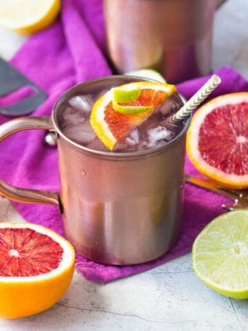 Winter Citrus Moscow Mule in a copper Moscow mule mug on a purple napkin surrounded by pink grapefruit and other citrus