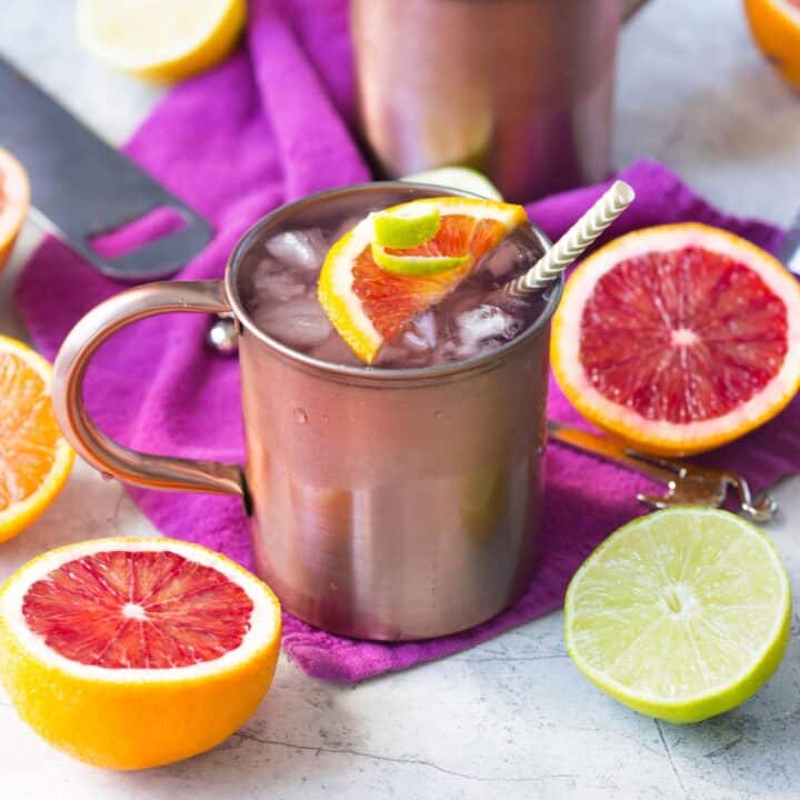 Winter Citrus Moscow Mule in a copper Moscow mule mug on a purple napkin surrounded by pink grapefruit and other citrus