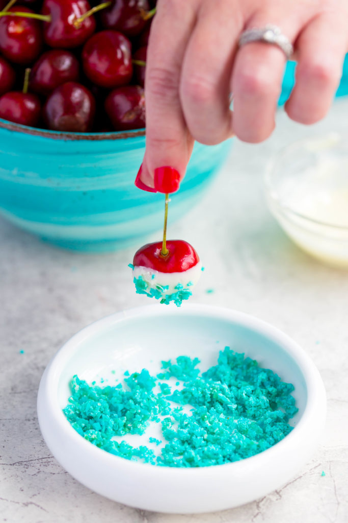 4th of July Desserts: Fresh white chocolate covered cherry being dipped in bowl of blue pop rocks