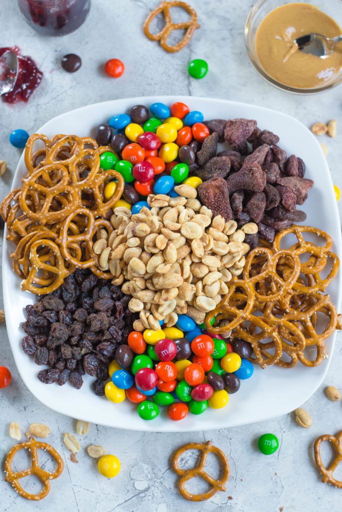 Peanut Butter and Jelly Trail Mix Ingredients: Peanut butter m&ms, dried strawberries, pretzels, raisins, roasted peanuts on a white plate