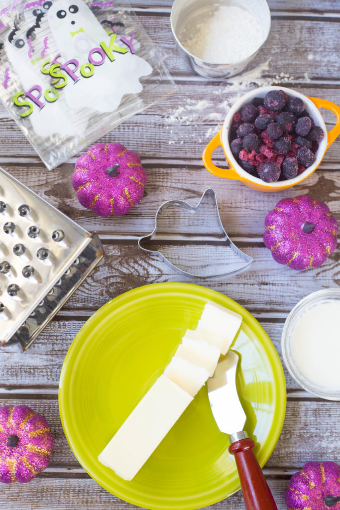 Overhead photograph of butter, cheese grater, purple pumpkins, frozen blueberries, and spooky Halloween bags on brown wood