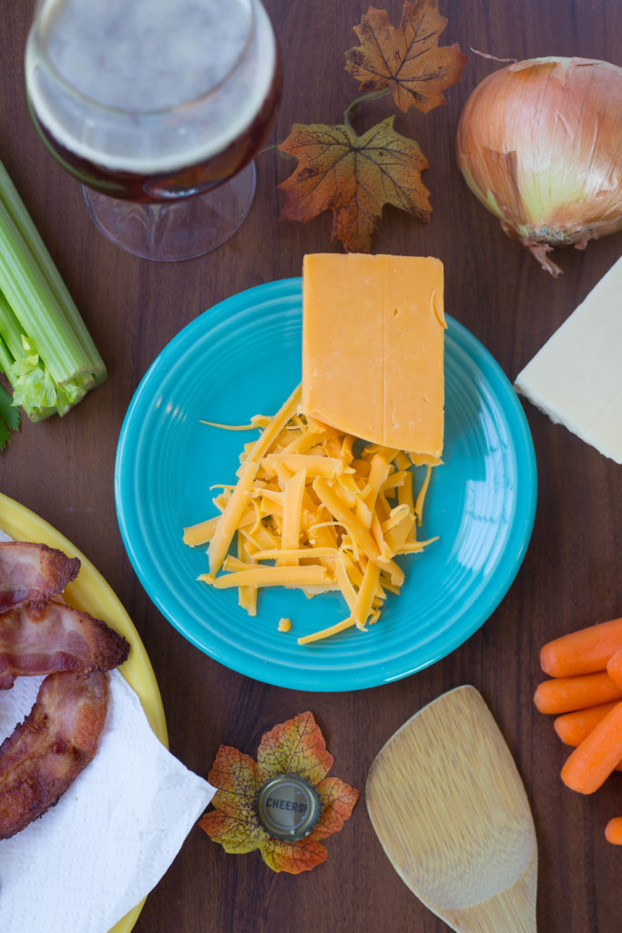 Block of orange cheddar cheese half grated on a plate surrounded by Octoberfest Beer Cheese Soup ingredients