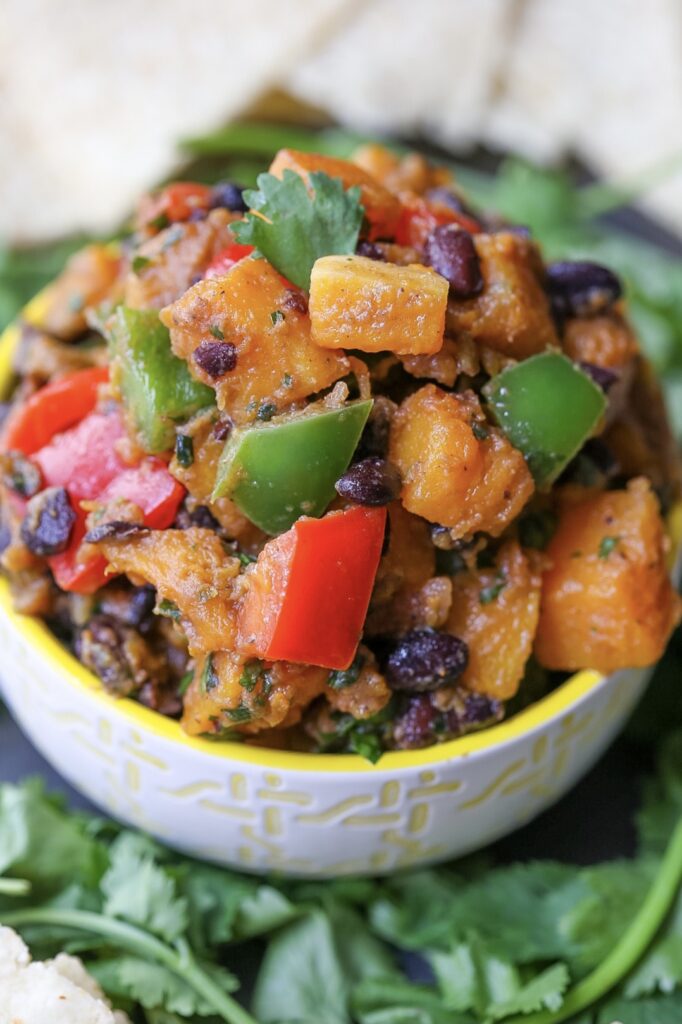 Chunks or roasted butternut squash, red and green bell peppers, and black beans, in a small bowl with yellow trim. 