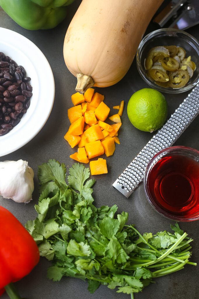 All of the ingredients for roasted butternut squash and black bean salsa ingredients: whole butternut squash, fresh limes, black beans, red wine vinegar, fresh bell peppers, fresh garlic. 