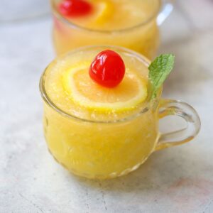 slushy pineapple party punch in small punch glass with lemon slice, mint leaf, and cherry on top