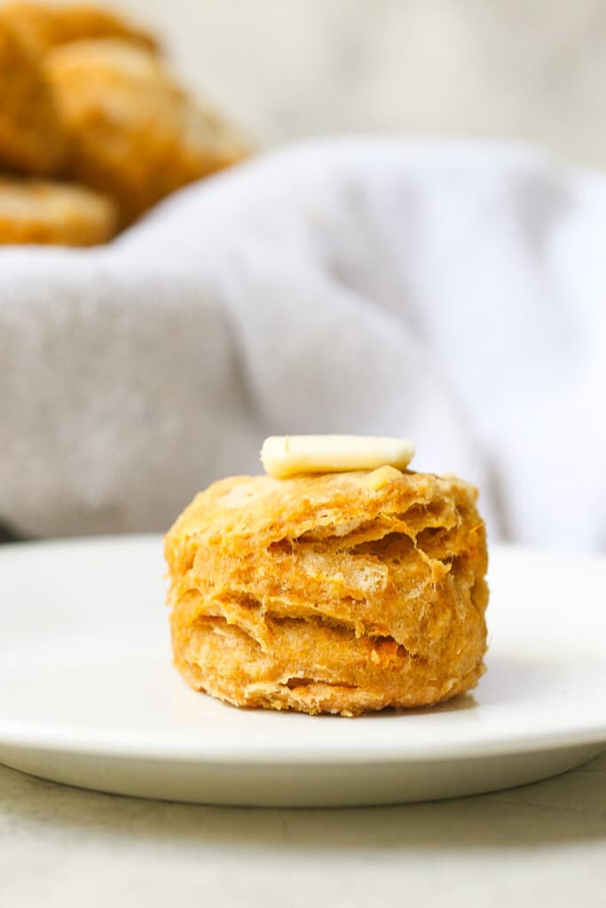 Close up of sweet potato biscuits with a pat of butter on top with a basket of biscuit in the background