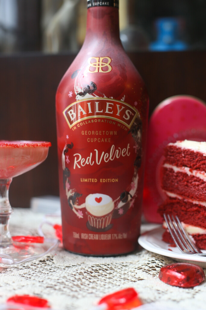 A bottle of Baileys red velvet cupcake Irish Cream liqueur on a lace covered table with a slice of red velvet cake on one side, and a red velvet martini on the other