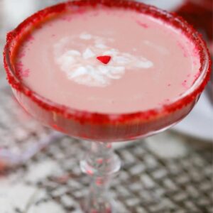 Red Velvet Martini in a glass with a red sugar rim, a drizzle of white cream, and a single heart sprinkle in the center