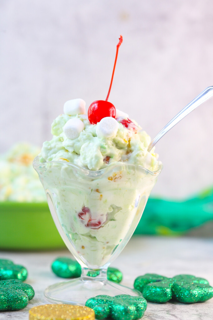 Ambrosia Watergate Salad with coconut and a cherry on top in an ice cream sundae glass surrounded by green decor