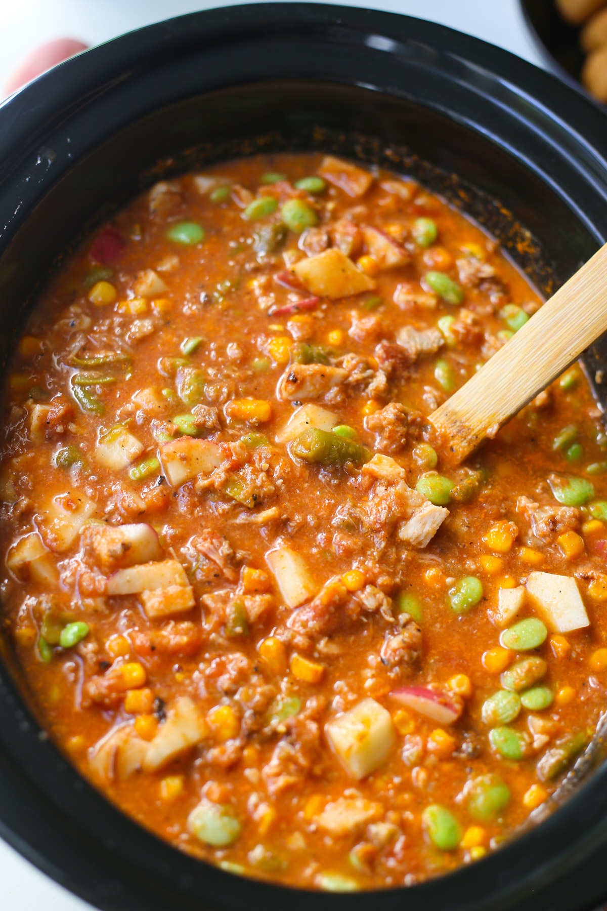 Brunswick Stew ingredients in a black crockpot with a wooden spoon handle sticking out
