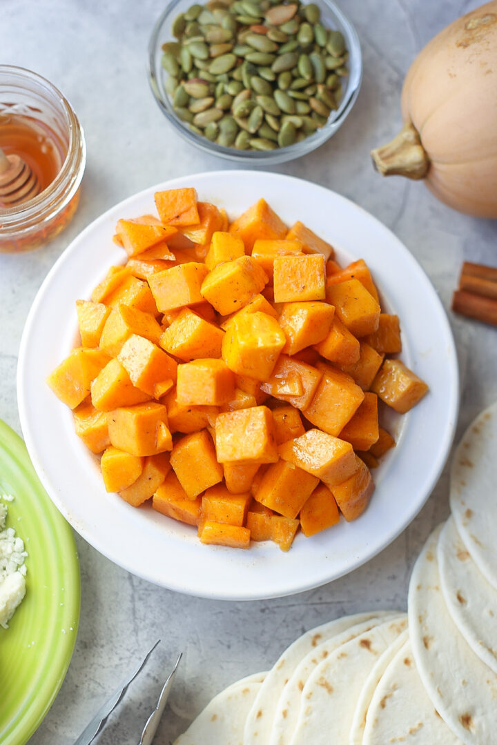 Ingredients for butternut squash tacos: cubed butternut squash in a white bowl, green pumpkin seeds, honey, four tortillas